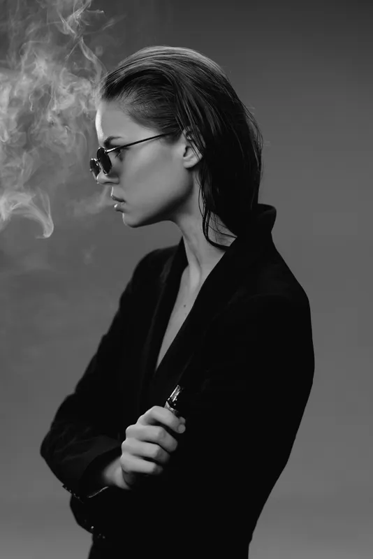 Stylish woman vaping in black and white with glasses and dark clothing, exhaling smoke in moody atmosphere.