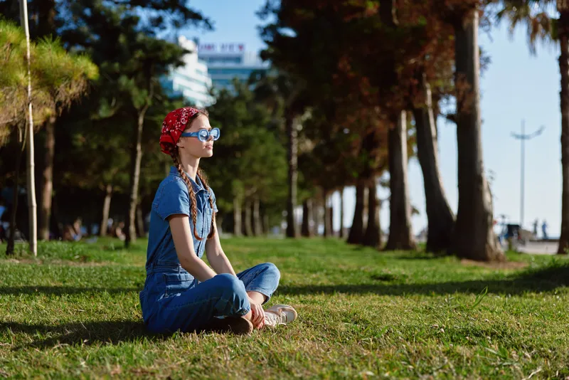 Lifestyle woman in modern boho-western denim street style sitting cross-legged on grass, wearing sunglasses and a red headband in film color outdoor setting.