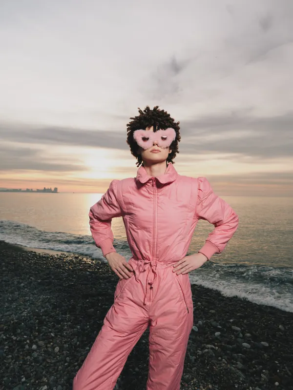 A woman on a rocky beach wears a pink jumpsuit and whimsical mask, posing confidently at sunset by the water, creating a bold, mysterious fashion moment.