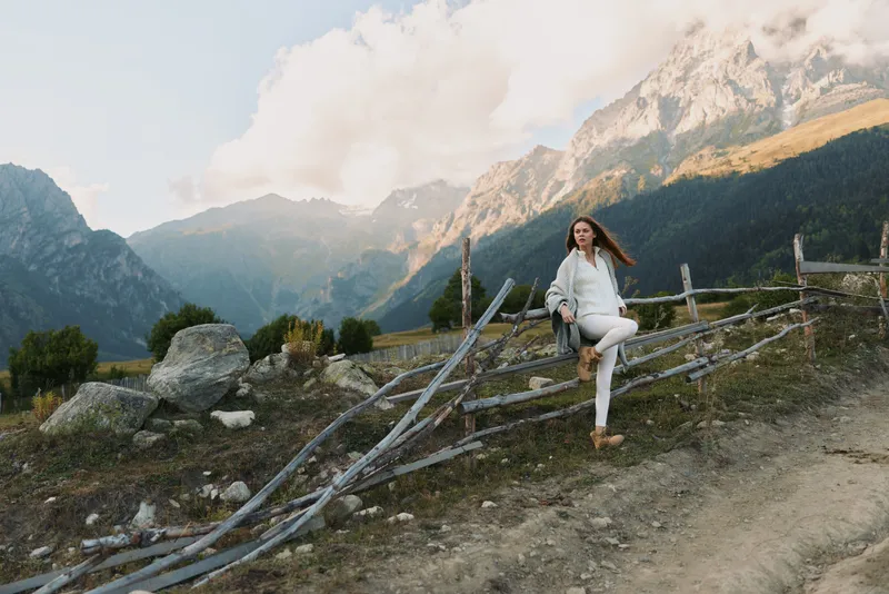 Woman lifestyle outdoors walking on mountain trail wearing white jacket and pants wooden fence rocky path scenic alpine landscape cloudy sky