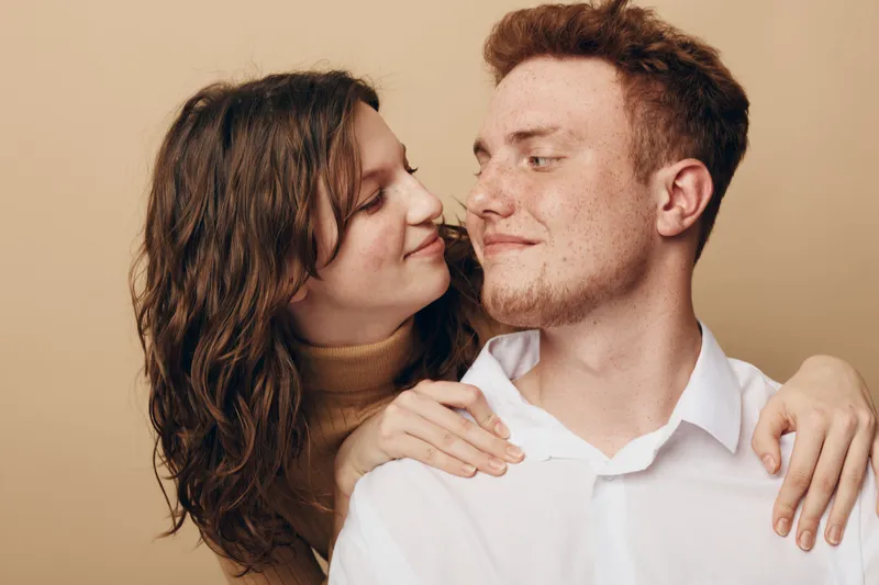 Young couple embraces tenderly, woman with curly hair smiles at man with short hair. Intimate moment of affection and love captured on neutral beige background in soft natural light.