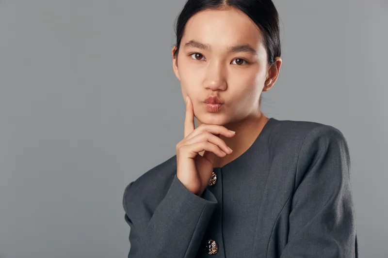Young woman with thoughtful expression and hand on chin wearing gray blazer looking at camera in studio. Portrait, contemplation and fashion with neutral background for professional concept.