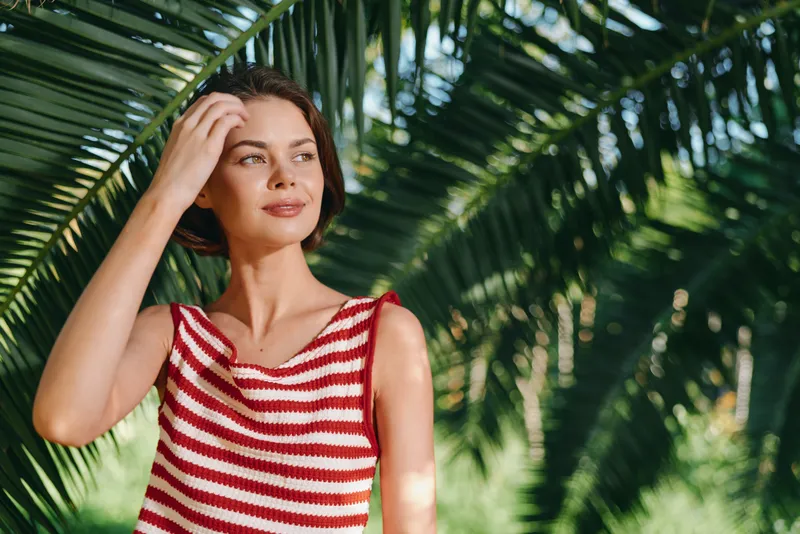 A thoughtful woman stands beside lush palm leaves, wearing a red and white striped top. She gazes into the distance with calm, sunlit confidence and graceful poise.