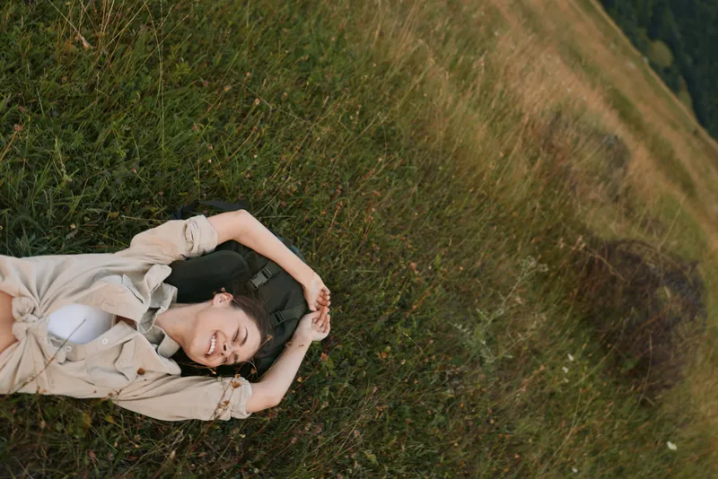 Young woman rests on grass in a sunlit meadow, eyes closed, wearing a light beige shirt, conveying calm relaxation and connection with nature.