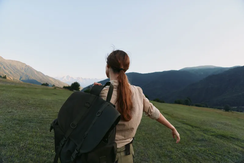 Woman with a backpack standing in a grassy valley facing distant mountains embracing outdoor life and exploration and a sense of freedom under wide open skies Natural light enhances a calm
