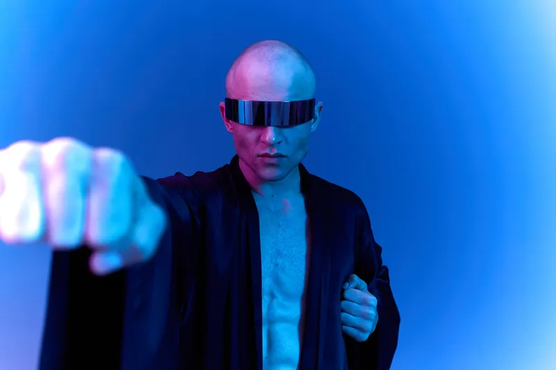 Futuristic man in a dark robe with sunglasses, striking a powerful pose, showcasing strength and confidence in a vibrant blue light. Black and white image.