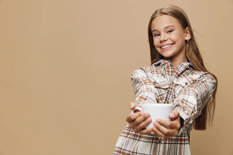 Smiling young girl holding white cup with both hands wearing plaid shirt with long hair on beige background. Friendly child offering drink in studio with happiness and casual pose.