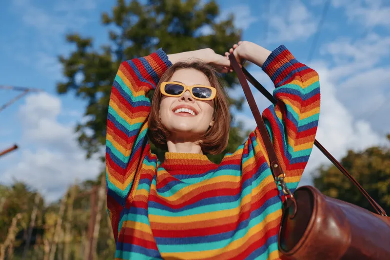Woman smile rainbow sweater sunglasses bag outdoors happy portrait of a young adult in colorful knitwear, casual fashion and carefree mood enjoying a sunny day with blue sky background