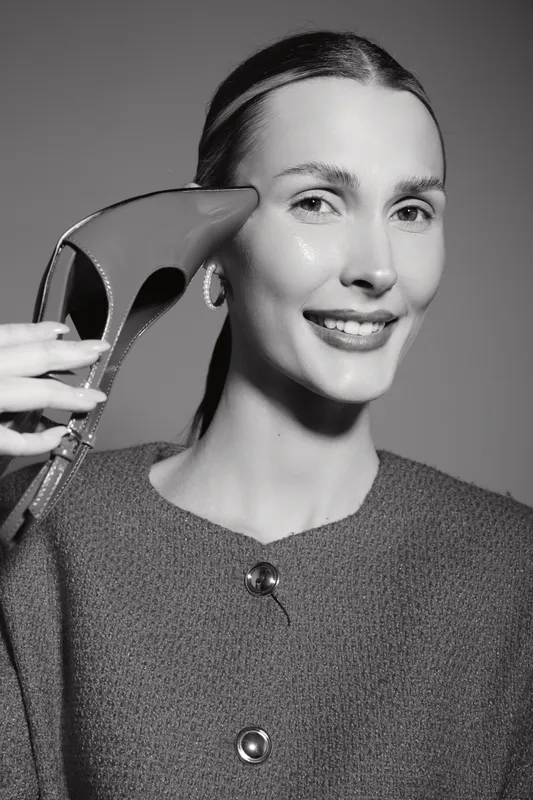 Smiling white woman with fair skin holds high heel shoe near face, wearing knitted sweater and earrings in black and white portrait.