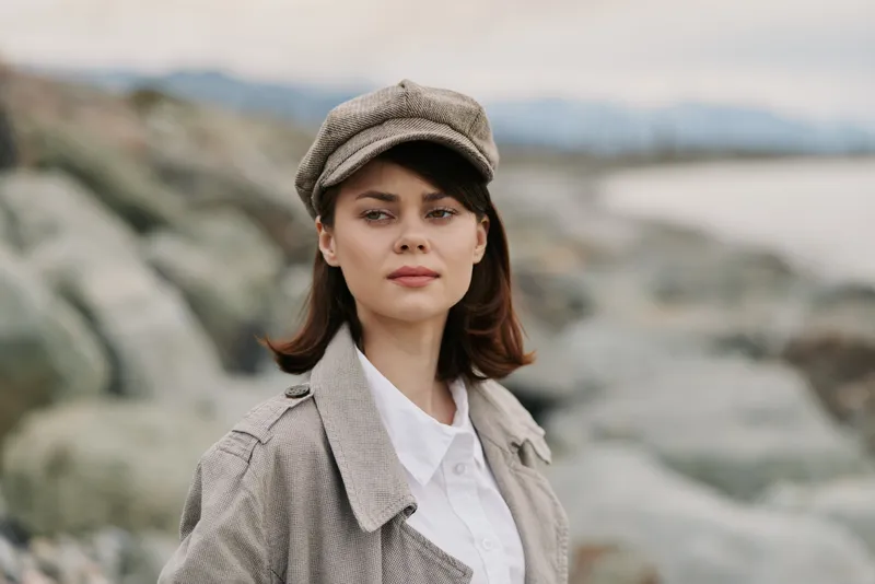 Young woman in tweed coat and cap stands outdoors near rocky coast with calm sea and cloudy sky. Fashion portrait with natural makeup and confident expression.