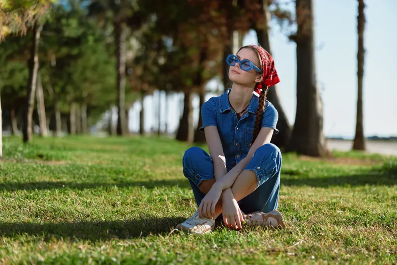 Lifestyle woman in modern boho-western denim street style sitting on grass with sunglasses and red bandana, film color tones capturing a relaxed outdoor vibe.