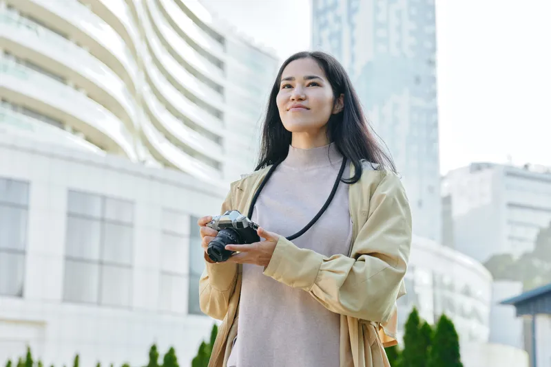 Camera in hand, woman explores urban cityscape with a relaxed outdoor lifestyle look, capturing candid moments and modern architecture during daytime in a bright courtyard with soft light