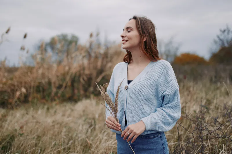 A young woman stands in a windswept field holding dried grasses, captured in an authentic, natural moment that emphasizes genuine emotion, relaxed posture and soft autumn light.