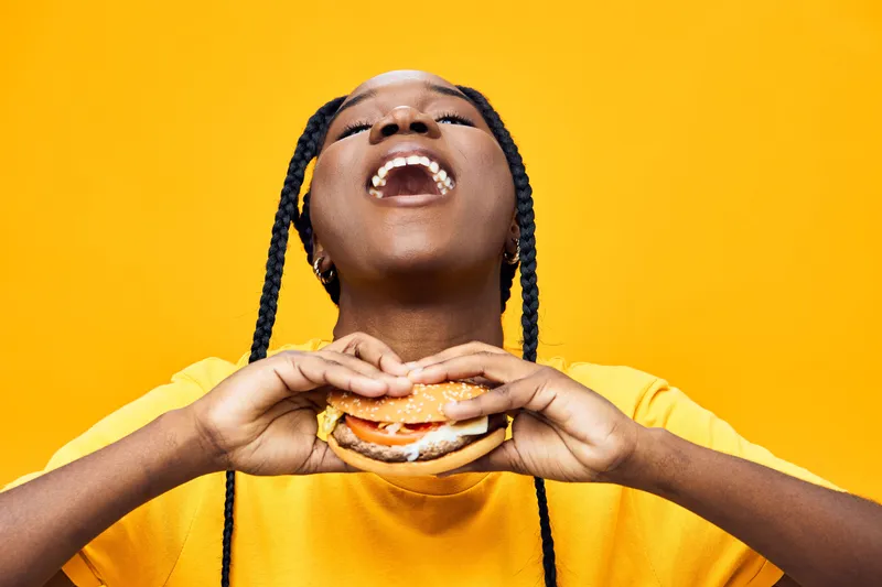 Happy Black woman enjoying a delicious burger against a bright yellow background, showcasing joy and food culture, perfect for healthy eating concepts