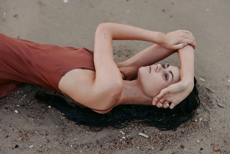 Portrait of a woman lying on sunlit sand in a rust dress, arms raised and crossed above her head, with a tranquil, contemplative expression and soft beach light.