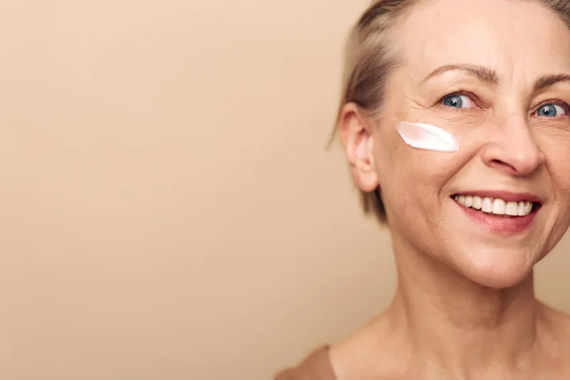 Smiling middle aged woman with blue eyes applies white face cream on cheek. Close-up on beige background. Concept for skincare, anti aging, beauty, wellness, and natural cosmetics.