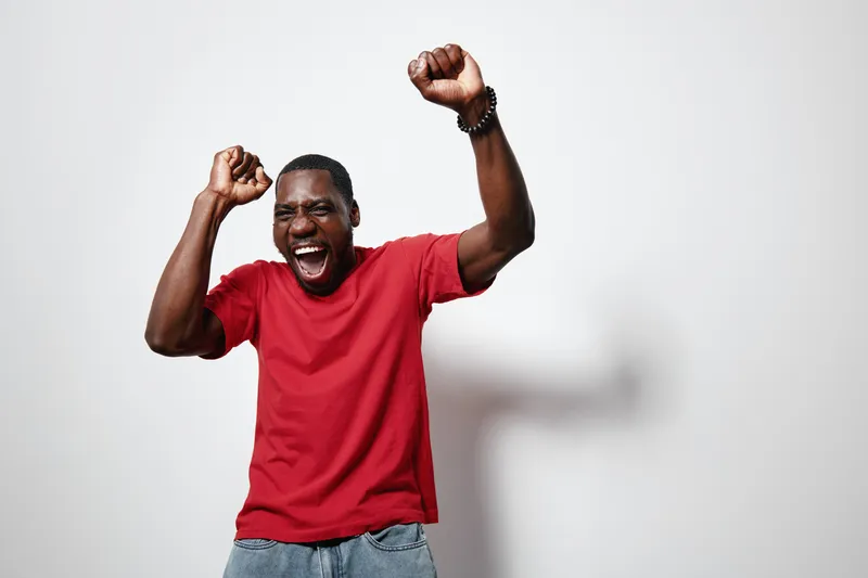 African American man with dark skin wearing red t shirt and jeans celebrating with raised fists and joyful expression on white background in studio.