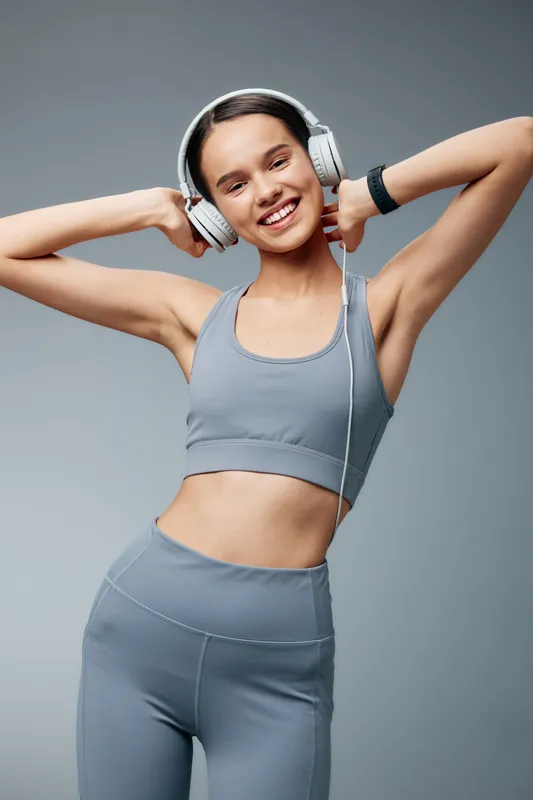 Woman fitness model wearing gray sportswear and headphones in a studio, posing with arms raised. Energetic workout vibe, active lifestyle, focus on fitness and music, bright lighting and clean