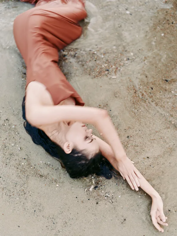 A solitary figure lies on sandy ground, arms extended, wearing a rust-colored dress, creating a quiet, contemplative moment captured from above with soft natural light.