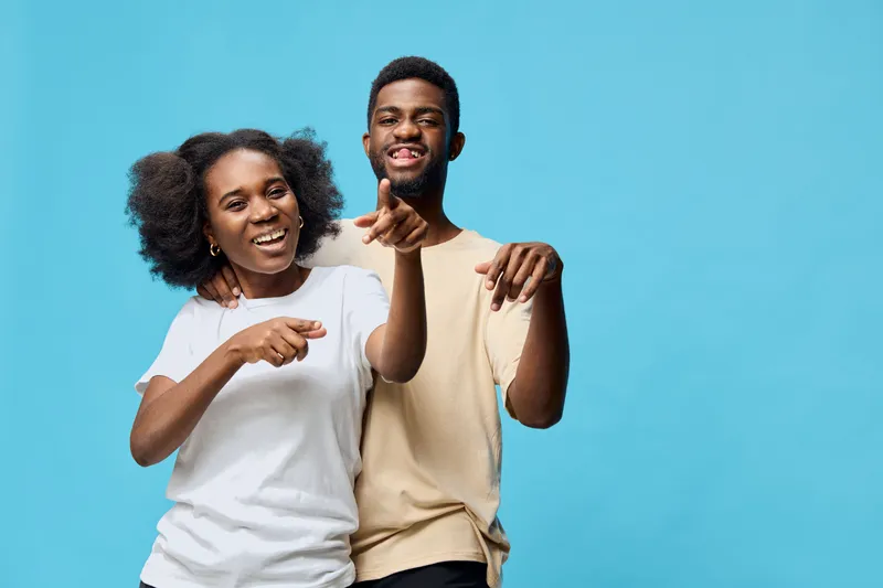 Happy young Black couple laughing and pointing at each other against a light blue background, showcasing joy and connection in a playful moment