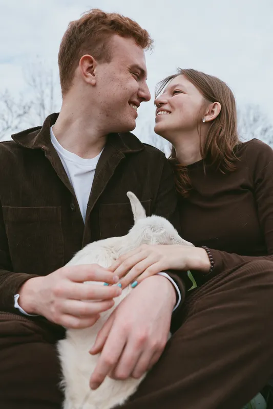 A couple sits close outdoors with a gentle goat kid between them, smiling and holding hands, capturing warm romance, affection, and family vibes in a casual lifestyle moment, content and intimate