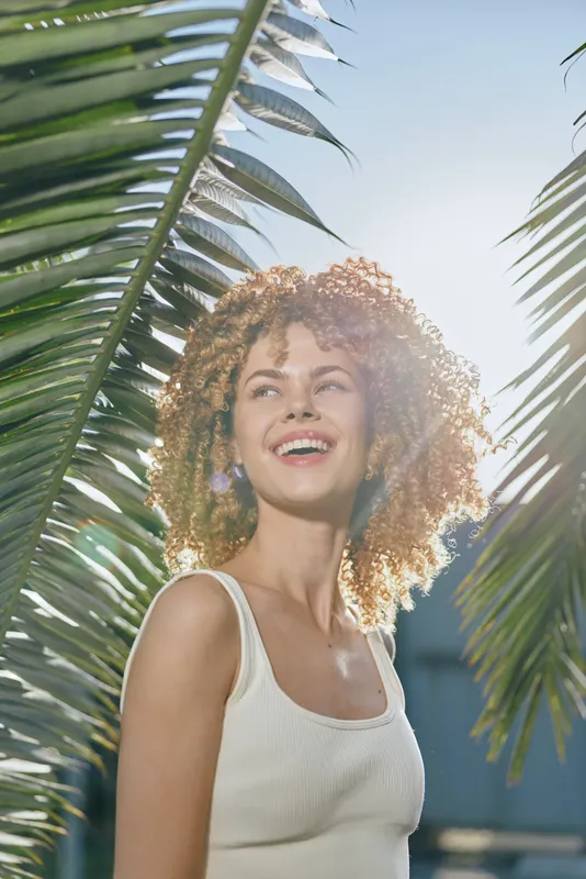Smiling woman with curly hair enjoys bright sunlight outdoors framed by green palm leaves, wearing a white sleeveless top and radiating natural joy in a tropical setting.