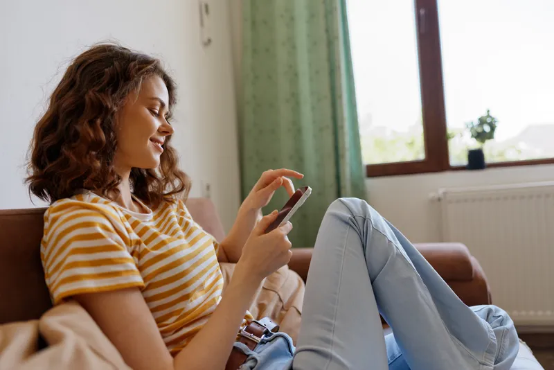 A woman sits on a sofa at home, scrolling on her smartphone with a relaxed expression. A bright living room scene captures casual lifestyle, comfort, and everyday digital connection.