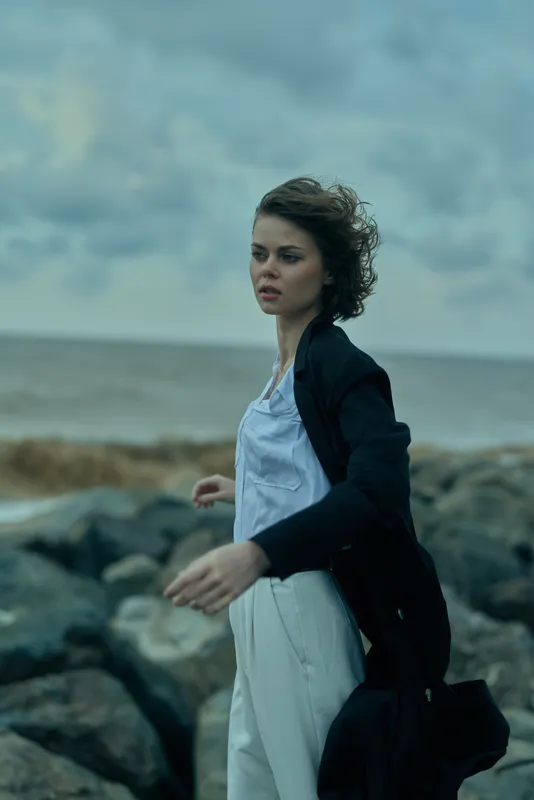 Windy coastal scene with a woman in a white dress and dark coat, walking on rocks, gaze toward the horizon as waves roll in, moody ocean backdrop and dynamic movement
