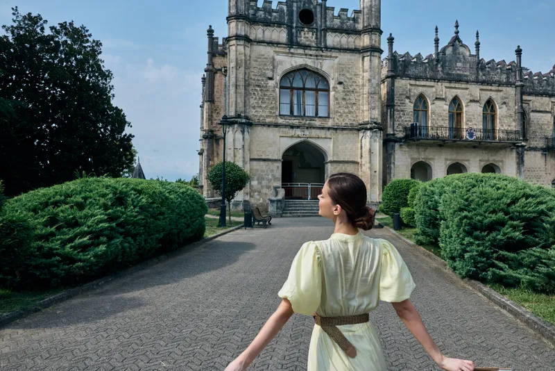 woman historic mansion garden architecture pathway dress backview authenticity inclusivity, female traveler in vintage pale dress walking toward ornate stone manor, serene scene