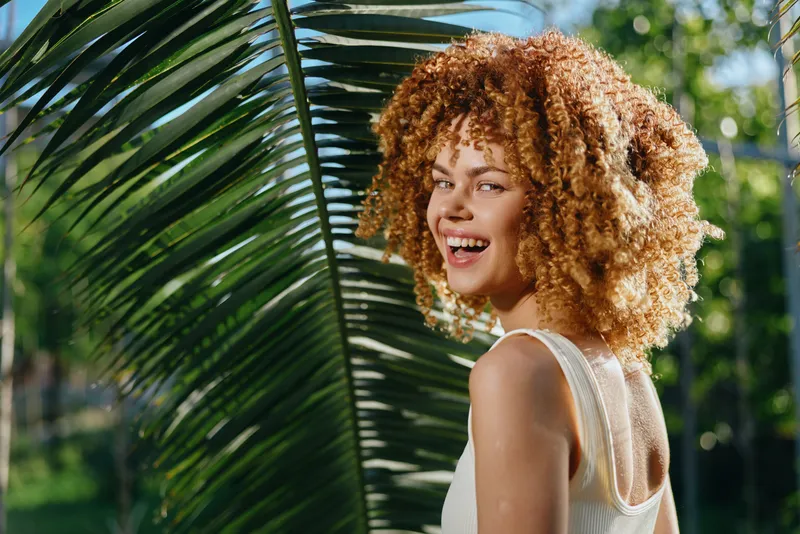 smiling woman with curly hair wearing white top, standing near green palm leaves in bright natural sunlight, vibrant and fresh atmosphere