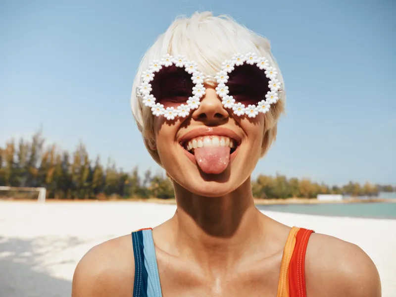 Playful portrait of a young woman at the beach wearing floral sunglasses, smiling with tongue out, sunlit sand and sea behind, bright summer mood.
