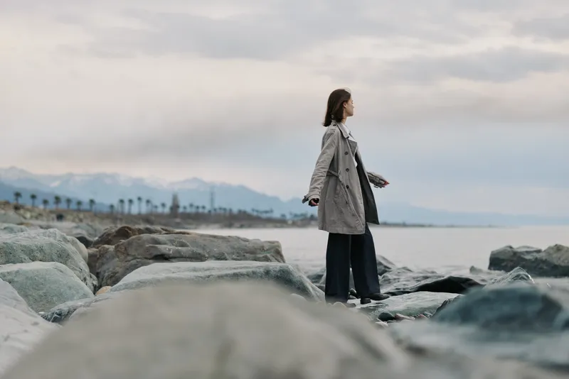 A woman in a long coat stands on rocky shore facing the calm sea, mountains in the distance under cloudy sky. Concept of solitude, nature, travel, and peaceful contemplation outdoors.