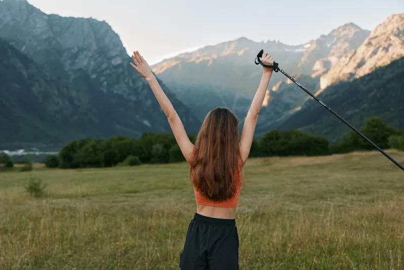 A person stands in a wide valley with towering mountains in the background, raising arms in triumph while holding a trekking pole, expressing joy, freedom, and adventure.
