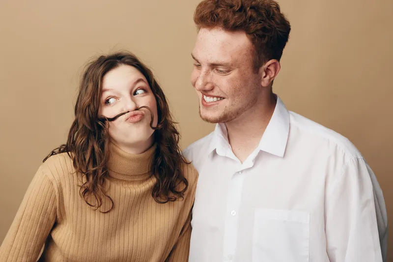Playful young woman with curly hair makes a mustache with her hair while smiling man in white shirt looks at her. Concept of fun, friendship, and lighthearted moments in casual studio setting.