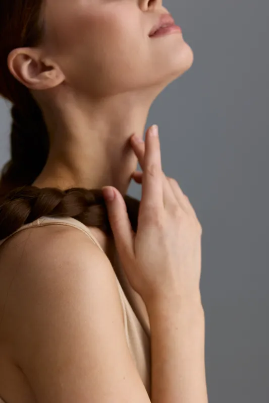Close-up of a woman’s neck and hand touching her throat, showcasing smooth skin and braided hair against a neutral gray background with soft lighting.