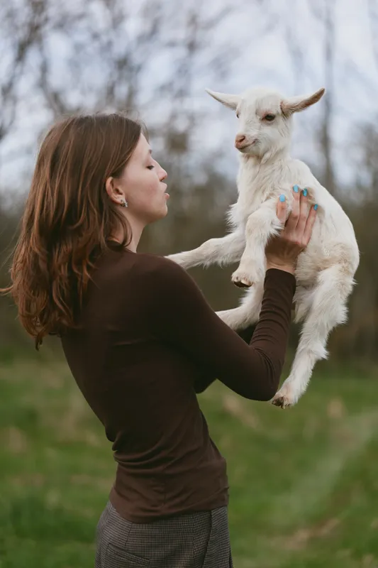 Young woman with long brown hair holds a white baby goat in her hands outdoors, enjoying a peaceful moment in a natural lifestyle setting during early spring.