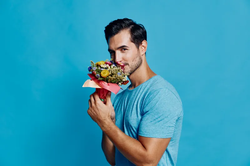 Handsome smiling man holding a colorful bouquet of flowers close to his face, showcasing his charming smile against a vibrant blue background. Floral concept.