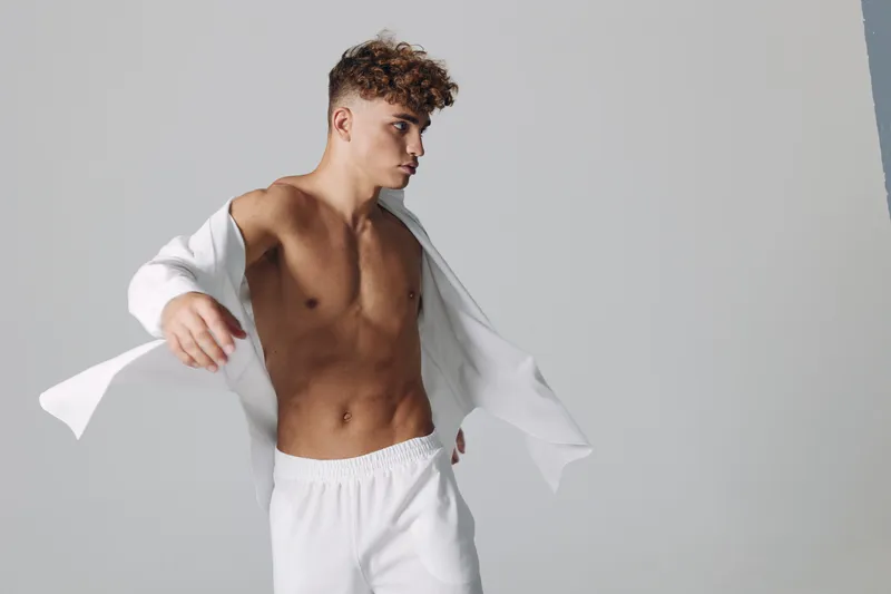 Young man with curly hair posing shirtless in white pants and open white shirt against plain background. Fitness, fashion and casual style with confident expression and toned body.