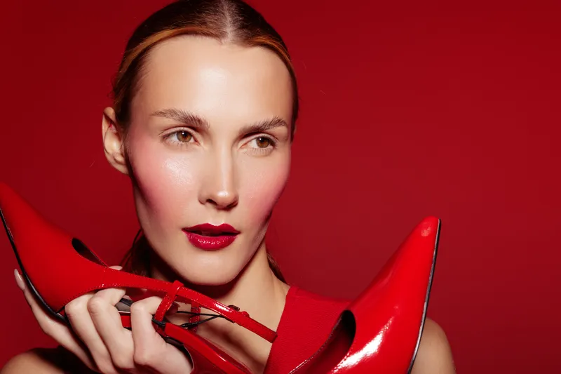 Close-up of a woman with glossy red lips holding shiny red high heels near her face. Red background and matching shoes create a bold fashion and beauty concept with vibrant colors.