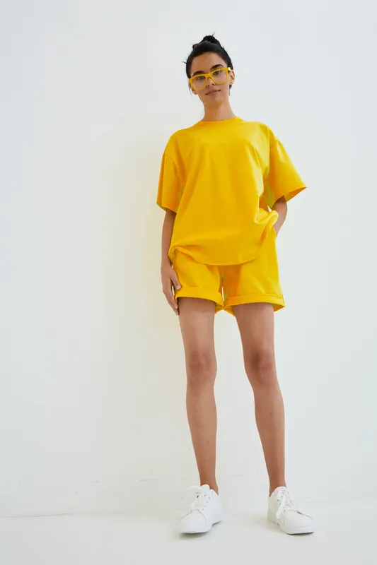 young woman wearing bright yellow casual outfit and sunglasses standing against white background with relaxed confident posture