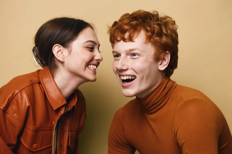 Young man and woman smiling and laughing together in casual clothing against beige background. Happy friends enjoying moment with joyful expressions and close connection.