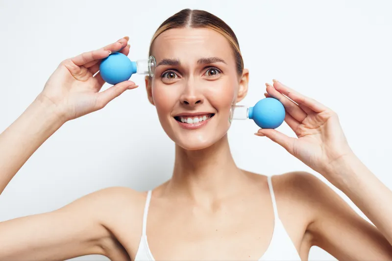 Young woman with clear skin holding blue facial suction cups near her face, smiling gently, clean background, beauty routine, skincare treatment, wellness concept