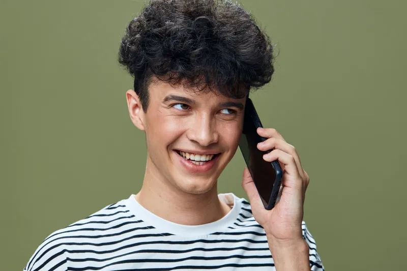 young man smiling and talking on phone indoors with striped shirt short curly hair casual style green background