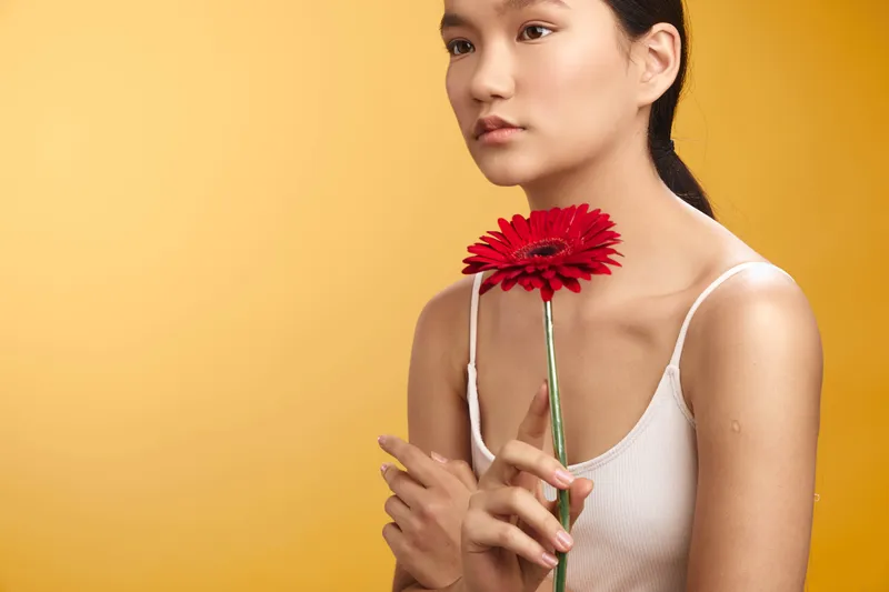 asian woman with dark hair holds a red flower against a warm yellow backdrop, gazing softly forward, conveying calm beauty and quiet elegance in a minimalist studio portrait.