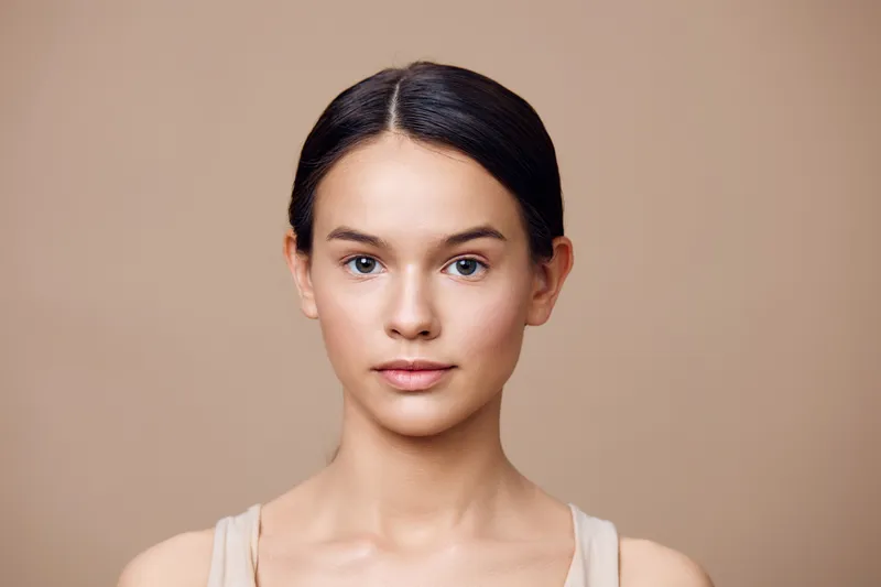 Young female model with natural beauty posing on a simple beige background, showcasing minimalistic style, calm emotions, and clean skin tone for beauty and wellness themes