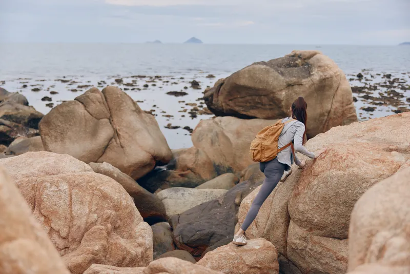 Young woman climbing rocky coastal terrain, showcasing adventure spirit and outdoor exploration, dressed in casual activewear with a backpack