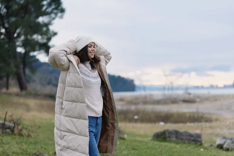 Woman outdoors wearing a long puffer coat in a windy lakeside landscape with fields and trees showing seasonal style and outdoor comfort for travel photos today