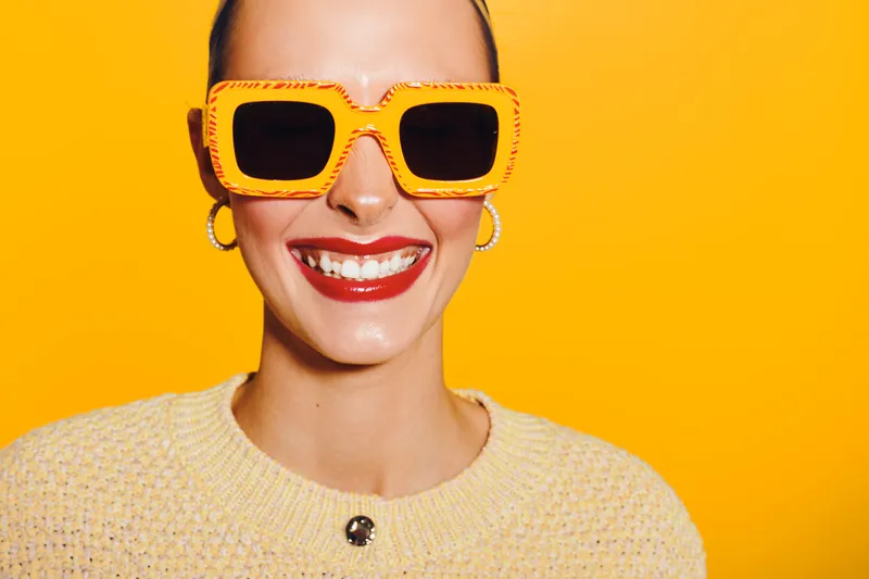 Smiling woman with bright lipstick and large yellow sunglasses poses against vibrant yellow background. Concept of fashion, summer style, and confident beauty portrait.