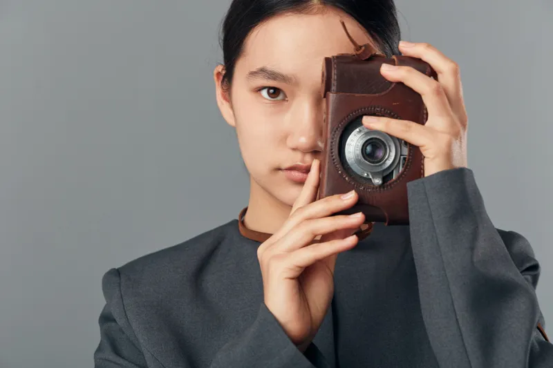 Young woman holding vintage camera in front of face with serious expression studio portrait. Female photographer with dark hair and gray outfit focusing on lens for retro photography concept.