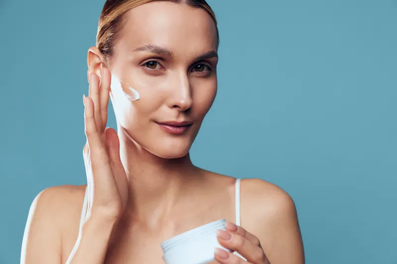 Woman skincare, face cream application, beauty close up, self care moment, she applies white cream, soft studio lighting, blue background, smooth healthy skin, calm mood, wellness, smiling